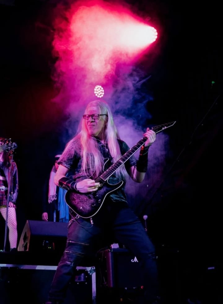 Hombre albino sosteniendo una guitarra eléctrica sobre un escenario iluminado con luces neón