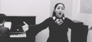 Mujer de vestido negro cantando con piano atras